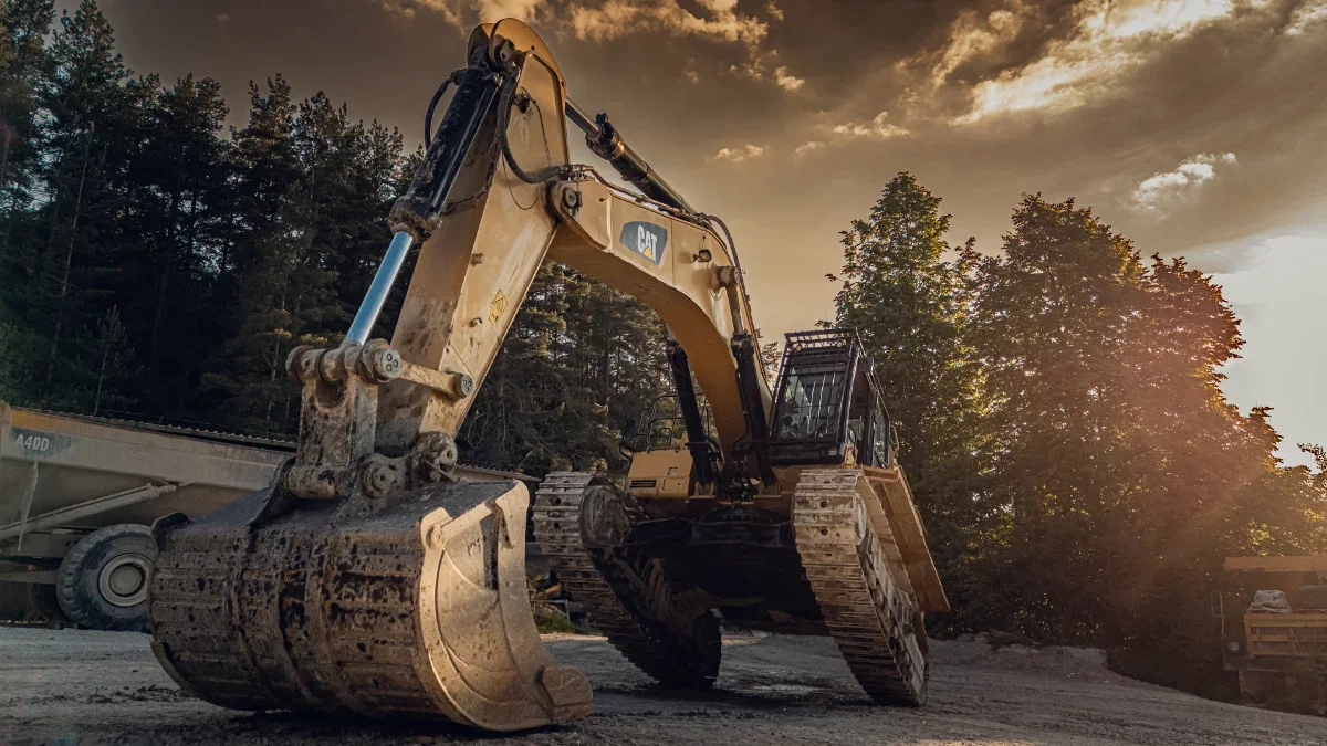 Large CAT crawler excavator with bucket lowered to the ground, tracked undercarriage visible, forest and dump truck in the background