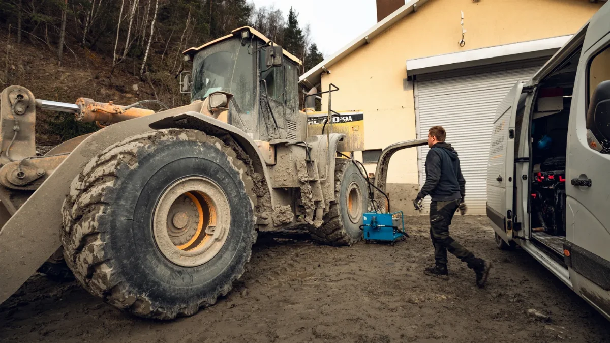 Mechanic walking from the WIBAKO service van to a muddy wheel loader, blue service equipment beside the machine