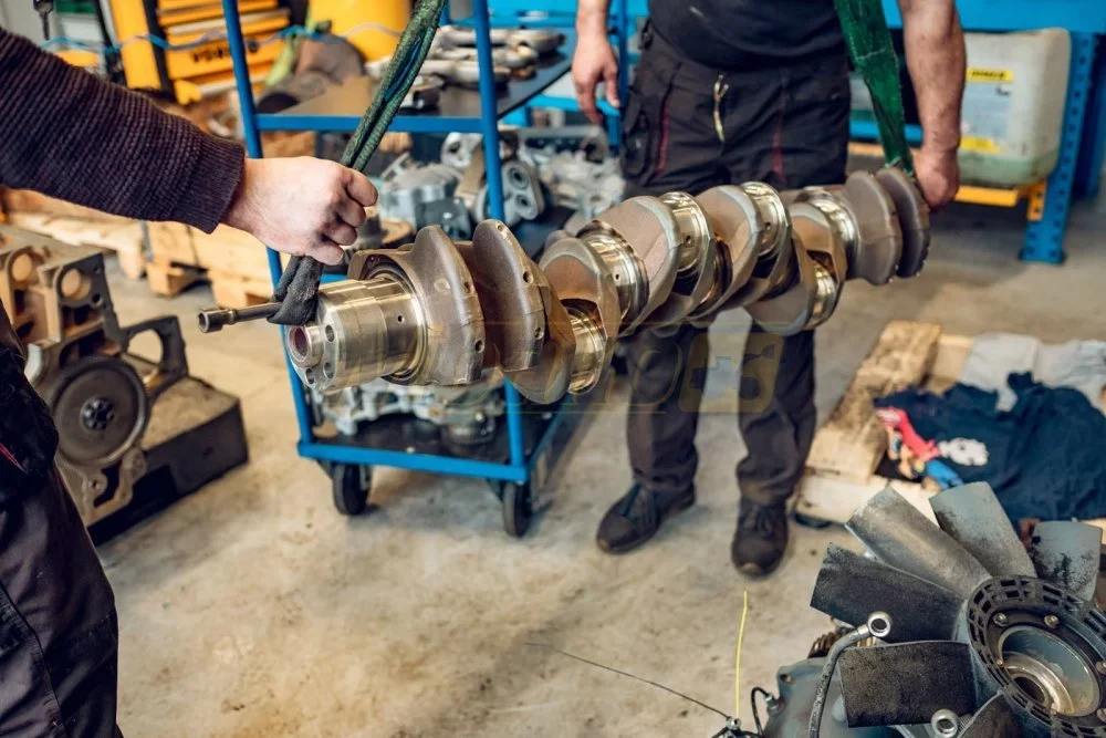 Mechanics carrying a diesel engine crankshaft on lifting straps in an engine rebuild workshop