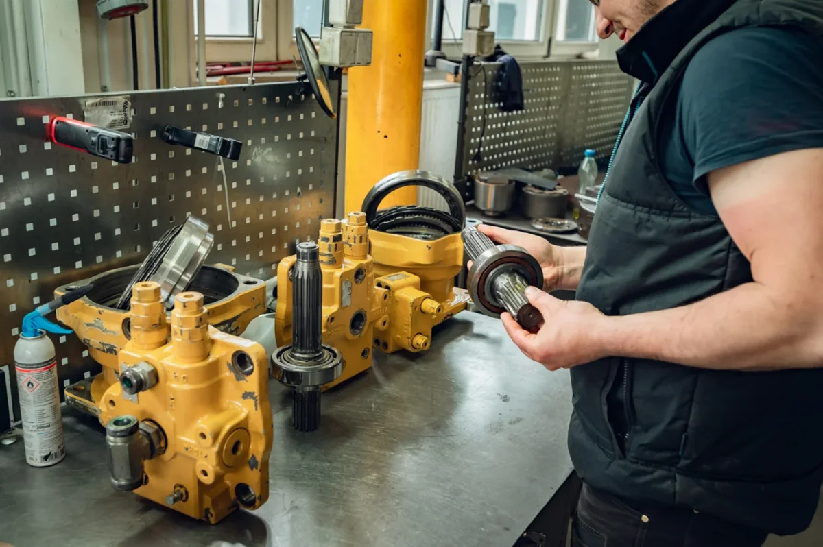 Mechanic inspecting a splined shaft with bearing next to a dismantled yellow hydraulic swing motor on a workbench