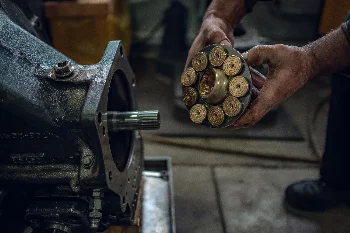 Mechanic's hands holding a piston set with slippers beside a hydraulic pump housing with splined shaft