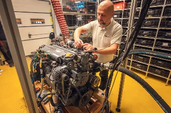 WIBAKO technician connecting lines to a diesel engine on a test stand, shelves with spare parts in the background