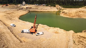 Hitachi long-reach excavator and service vans by a water-filled gravel pit, aerial drone view