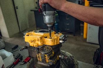 Mechanic using an impact wrench to unbolt a yellow hydraulic pump on a workshop bench