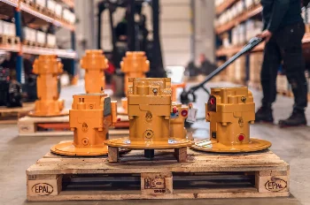 Yellow hydraulic swing motors on an EPAL pallet in a warehouse, worker with a pallet truck in background