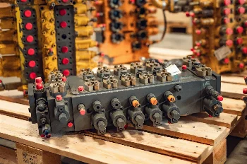 Hydraulic control valve block with capped ports on a pallet, yellow construction machine valves in background
