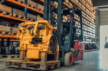 Linde E15 forklift carrying a yellow diesel engine on a pallet in a warehouse with parts racks